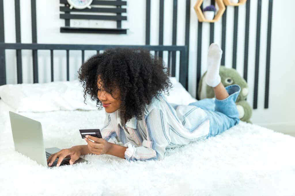 young woman laying down hold a credit card in her hand in front of a laptop